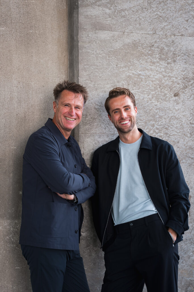 Twee mannen staan naast elkaar en glimlachen naar de camera - een echt Generatie-moment. De een, met kort bruin haar en een marineblauw jasje, slaat zijn armen over elkaar; de ander staat met teruggestoken haar, een licht overhemd en een zwart jasje tegen een betonnen muur.