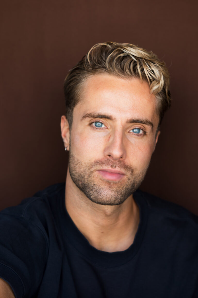 Een jonge man met een lichte huid, kort blond haar, blauwe ogen en licht gezichtshaar draagt een donker shirt en een kleine oorbel. Hij vertegenwoordigt de nieuwe generatie en kijkt met een neutrale uitdrukking naar de camera tegen een effen bruine achtergrond.